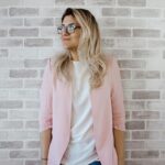 Elegant woman with glasses poses against a brick wall in a trendy pink blazer.